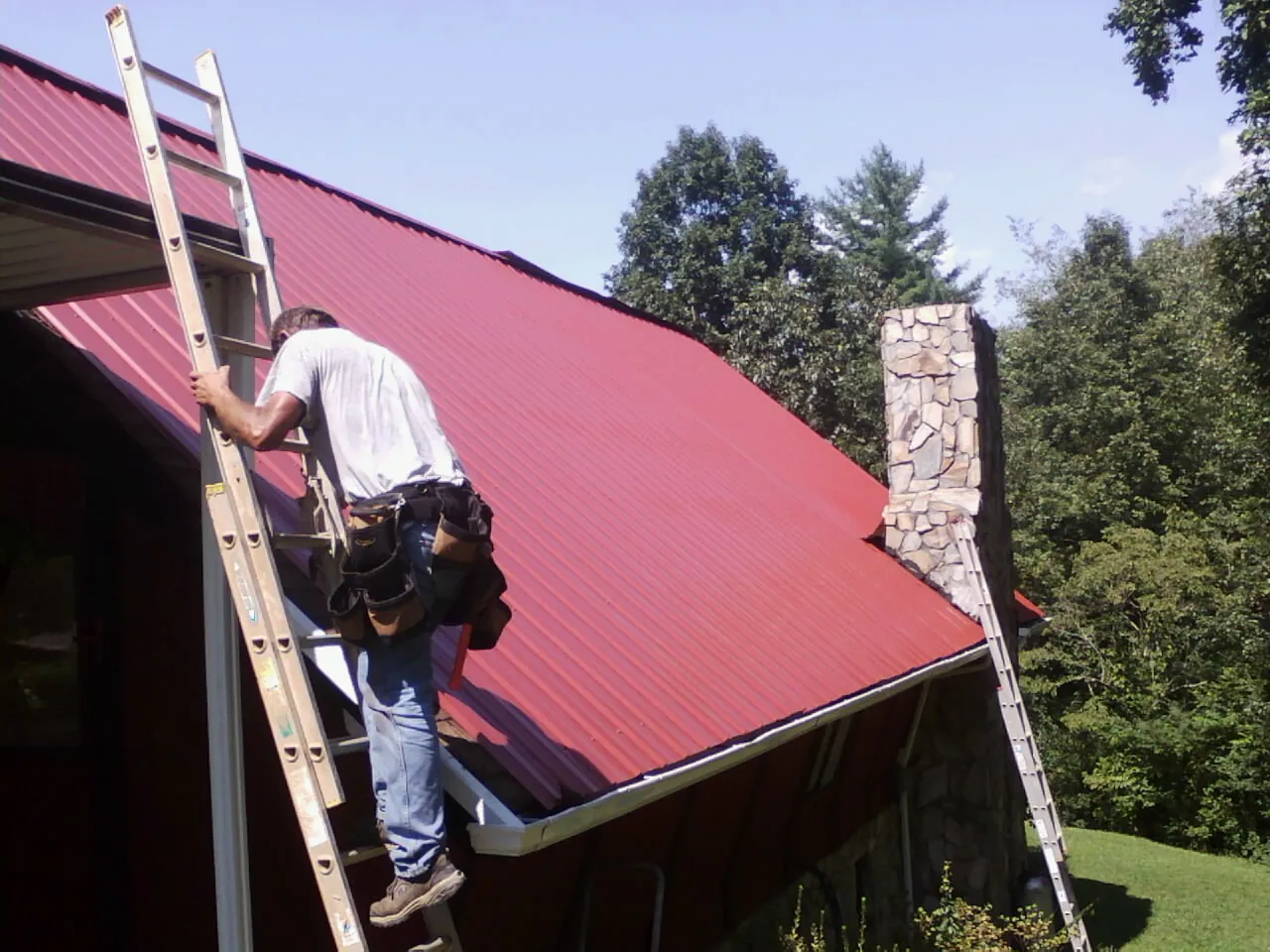 Roofer on a ladder beside a red metal roof and stone chimney — Soffit & Fascia Repair by Roofing Brothers in Oxbow